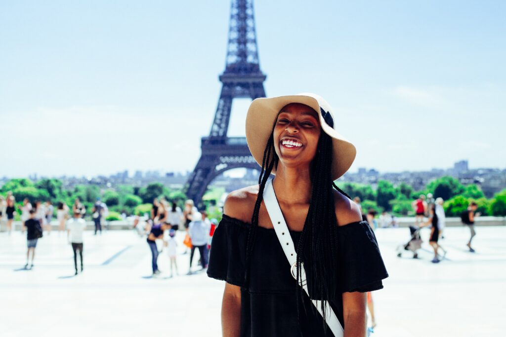 A young woman wearing an oversized sunhat smiles in front of the Eiffel tower. She is wearing an off the shoulder black dress and a white purse.