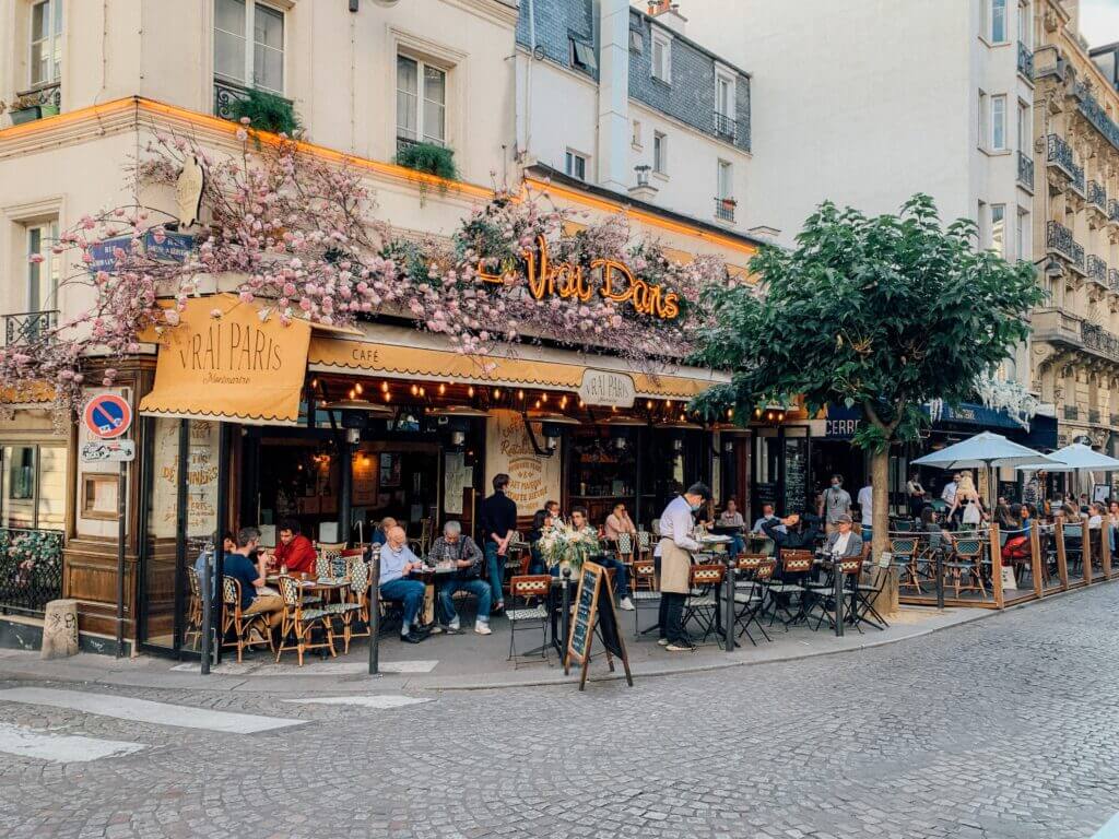 A Parisian street. The cafe Vrai Paris is in the foreground. Patrons of the cafe sit at small tables under a mustard yellow canopy. The cafe has a neon yellow sign displaying the name surrounded by pink flowers.