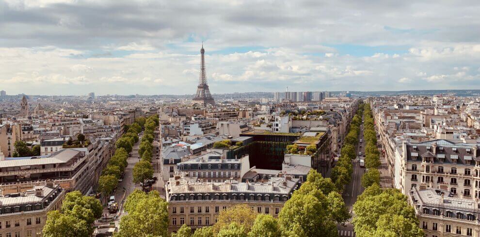 An aerial view of Paris. The Eiffel Tower sits in the background in the center of the image. Trees line the streets and the tops of buildings are visible. The sky is blue and cloudy.