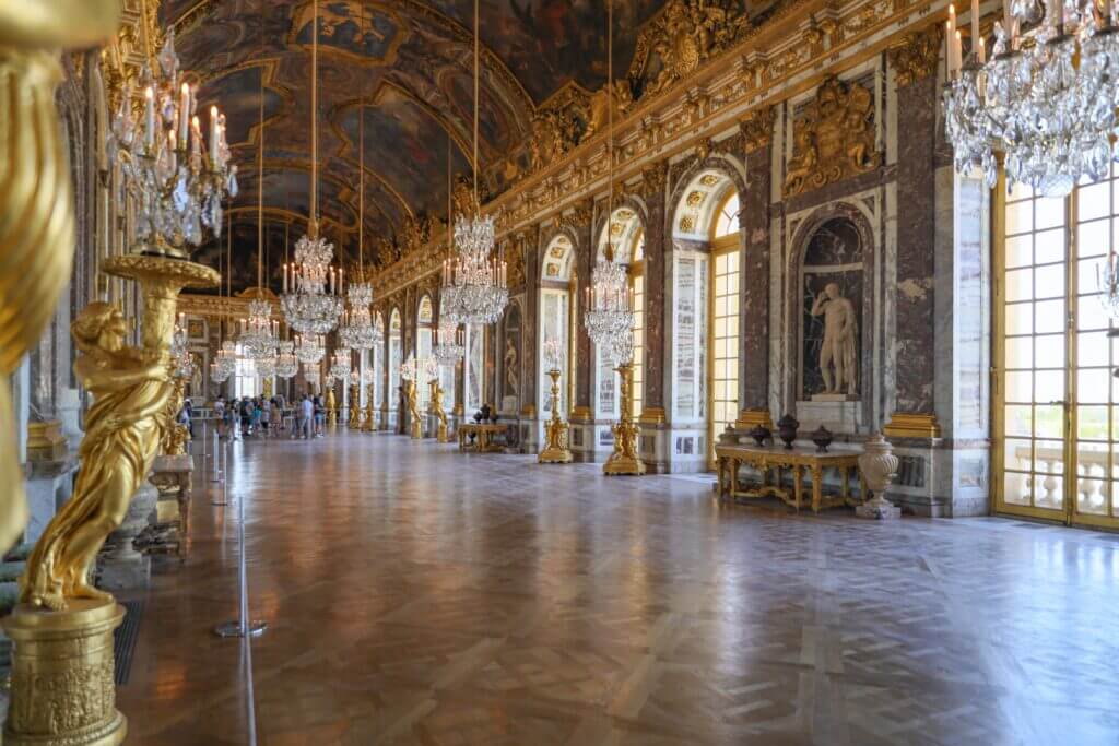 The hall of mirrors in the Chateaux Versailles. The entire hall is gold. The ceiling features intricate paintings in gold and blue. There are large floor to ceiling windows trimmed in gold lining the right side of the hall. Over 10 giant crystal chandeliers line the halls. There are gold statues lining the halls.
