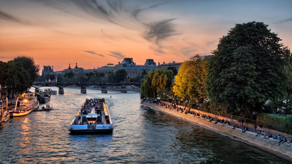 A riverboat cruises down the middle of the Siene river. The bank of the river is lined with lush green trees. The sky is orang and blue as the sun sets. The city skyline is visible in the background.