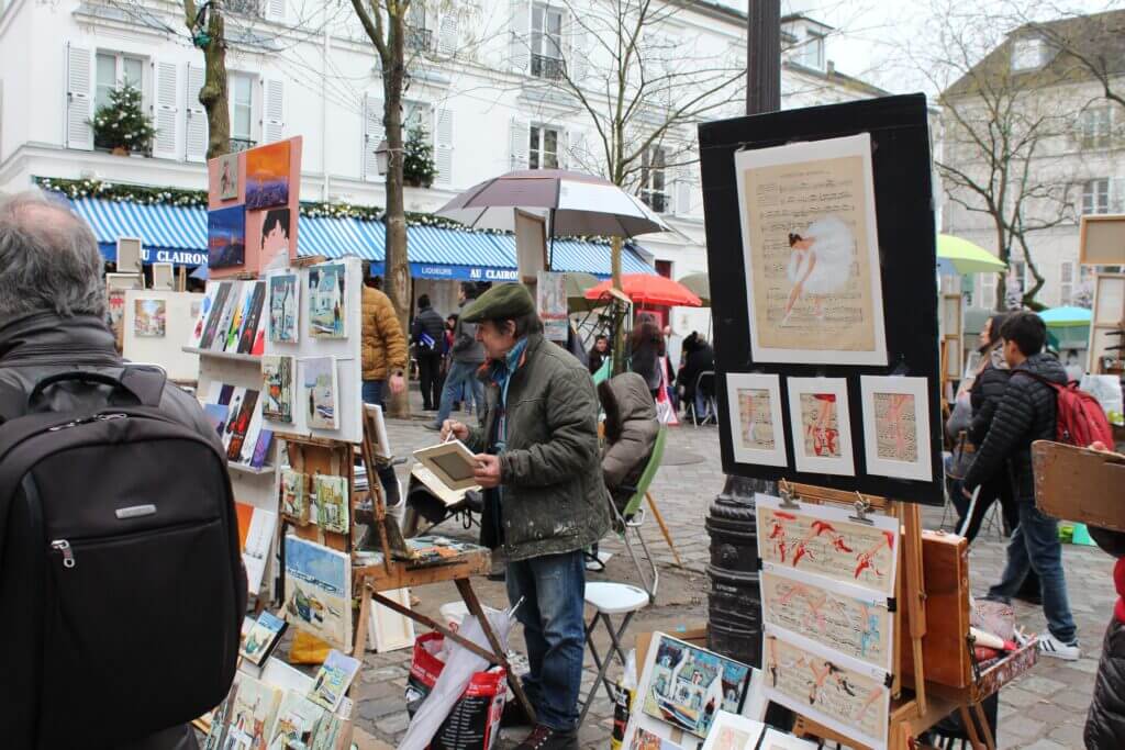Street artists in the Montmartre neighborhood selling their art. There is a man wearing am army green beret, army green coat, and blue jeans holding a small canvas and painting. His artwork displayed consists of many brightly colored landscapes. To the right of the man, there is another artist's set up. This artist is not pictured but is selling art of sheet music with ballerinas painted over it.