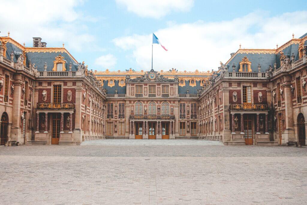 The front of Chateaux Versailles. The building is very extravagant. The exterior is a salmon color with blue roofing and gold trip. There is a French flag flying in the middle of the building. The sky is blue and cloudy.