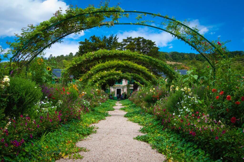 The entry way to Claude Monet's home in Giverny. There are 5 trellis arches covered in greenery growing over them. The dirft road leading to the pink house has lush greenery on either side. There are colorful flowers in bloom on the sides.